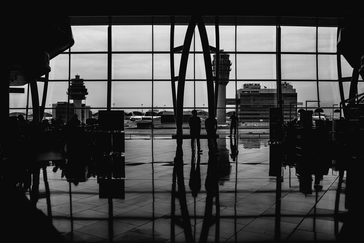Travellers silhouetted against the tall windows of an airport terminal, in black and white